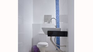 Baño con azulejos blancos, un lavamanos blanco, pared de ducha con azulejos azules y un cubo de basura morado.