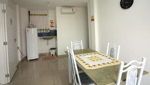 Comedor con mesa dispuesta para cuatro, sillas blancas, un reloj en la pared y cocina al fondo.