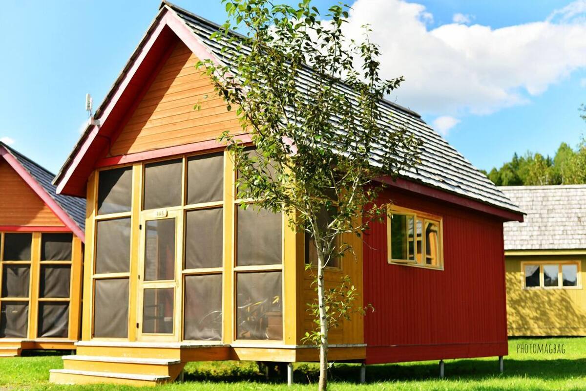 A red wooden house with large windows and a small tree in front.