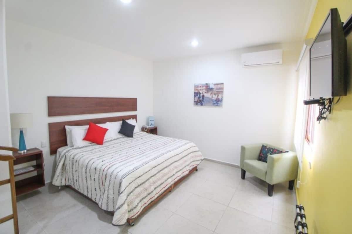 A bedroom with a bed, bedside tables, a chair, a TV, and an air conditioning unit.