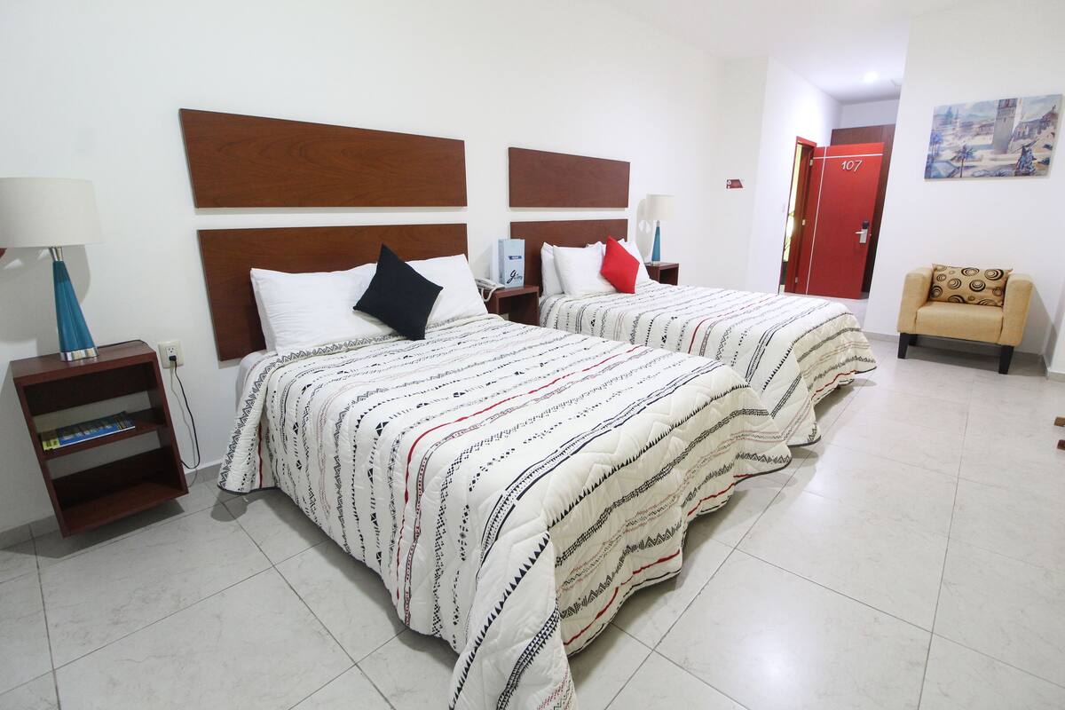 A hotel room with two beds, a small sofa, a nightstand with a lamp, and a bookshelf.