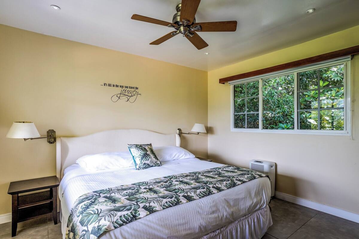 A bedroom with a bed, a ceiling fan, two bedside lamps, a window with a view of greenery, and a wall decal with text.