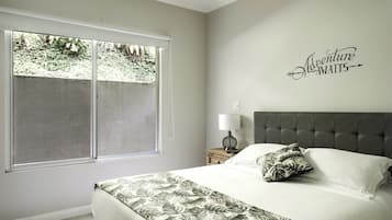 Ein Schlafzimmer mit großem Fenster, einem Bett mit floralem Überwurf, einem grau gepolsterten Kopfteil und einer Wand mit dem Schriftzug 'Adventure Awaits'.