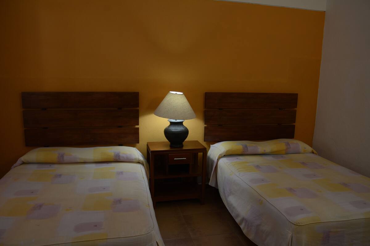 A room with two beds, each with a wooden headboard, a nightstand with a lamp, and a small drawer.