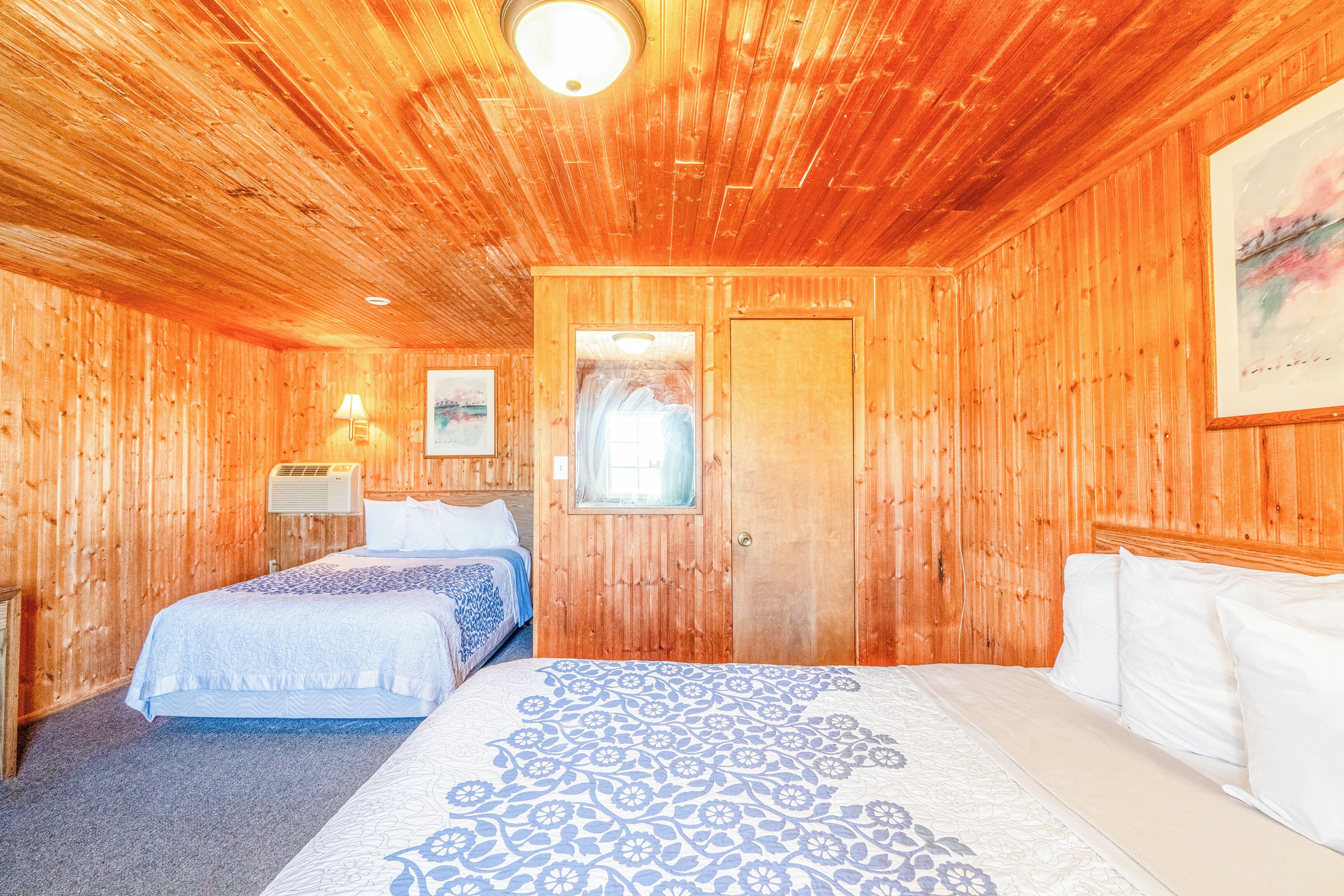 Habitación con paneles de madera, dos camas, una ventana y un cuadro en la pared.