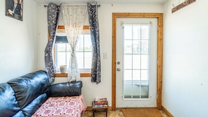 A room with a black leather sofa, a small table with a book, a window with curtains, and a wooden door. - Green Acre Motel by OYO Near Rush County Airport (La Crosse)