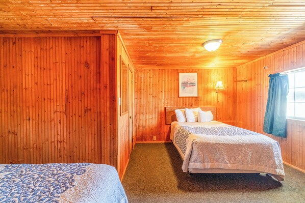 A wooden room with two beds, a nightstand, a wall-mounted lamp, and a window with curtains. - Green Acre Motel by OYO Near Rush County Airport (La Crosse)