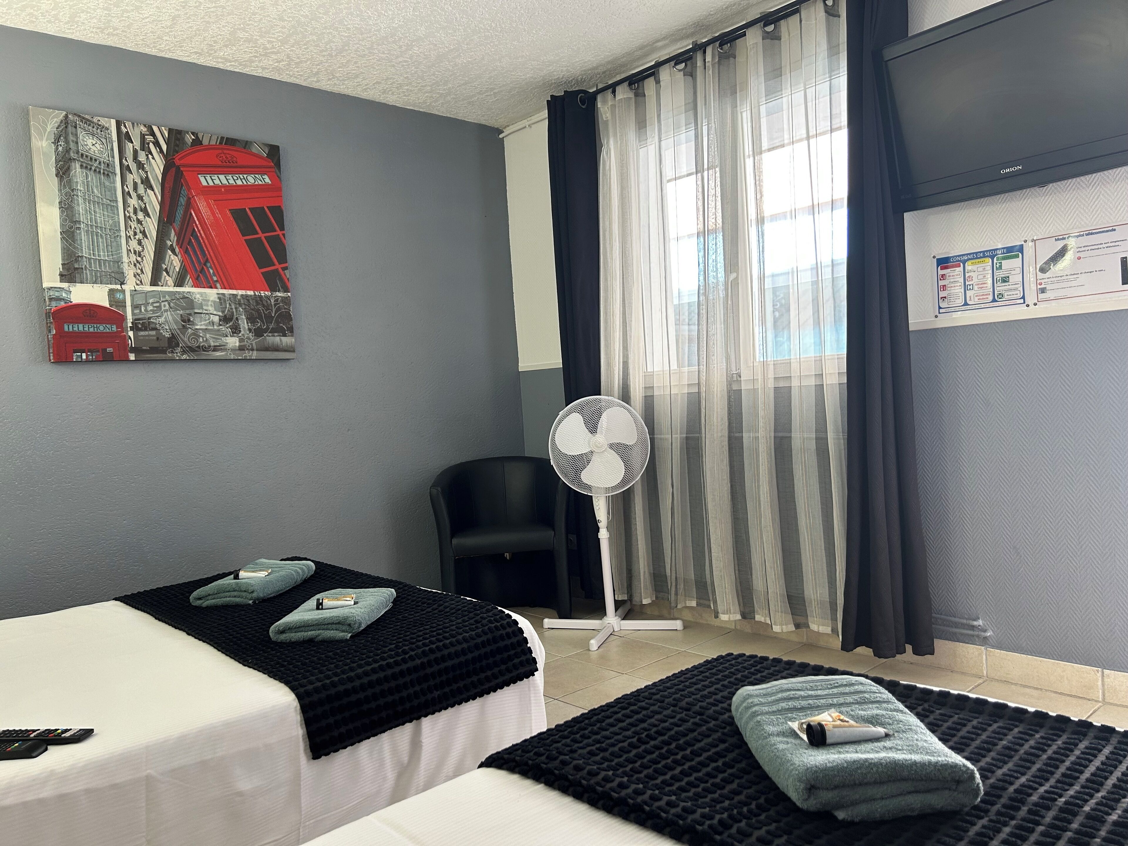 A hotel room with two beds, a fan, a red telephone booth artwork, and a window with sheer curtains.