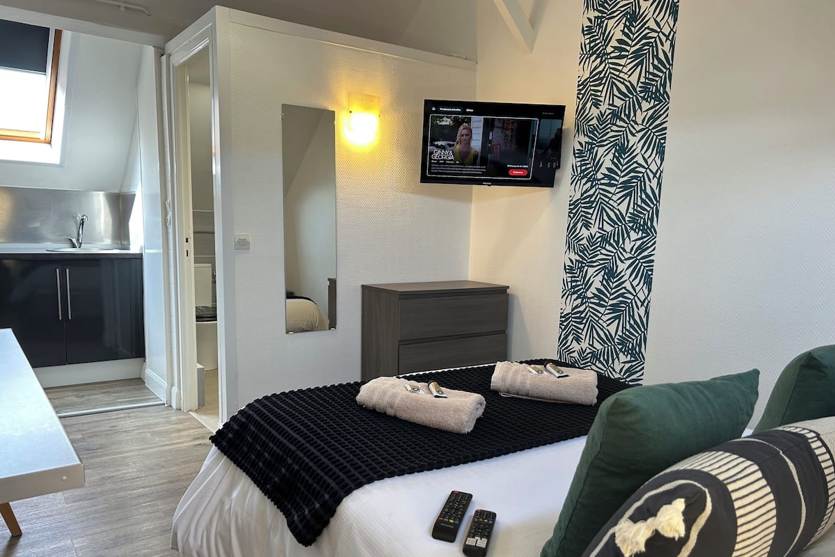 A modern hotel room with a bed, a TV mounted on the wall, a small desk, and a kitchenette in the background.