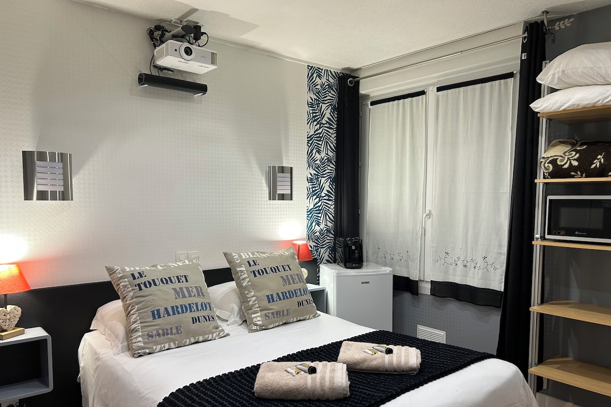 A hotel room with a bed, two bedside lamps, a projector, a small refrigerator, a black and white patterned curtain, and a shelf with pillows and a TV.