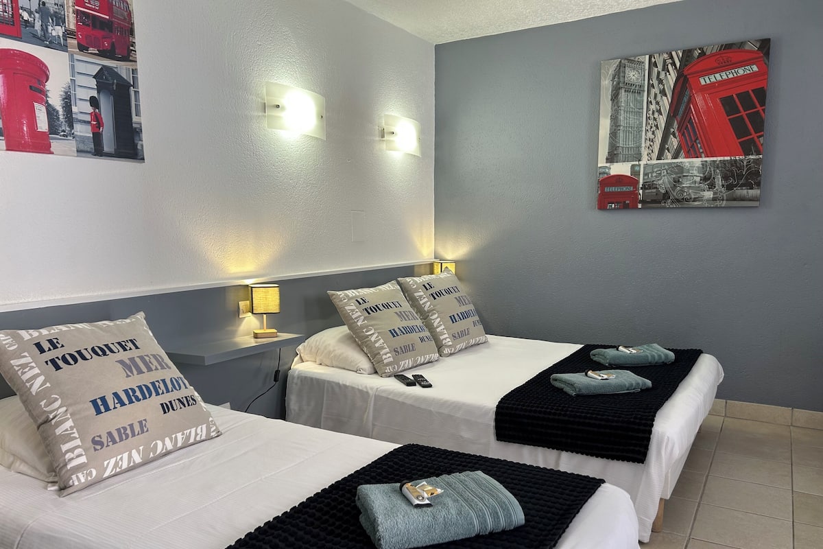 A hotel room with two beds, each with a white duvet and black throw blankets. There are wall-mounted light fixtures and a painting of a red telephone booth.