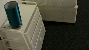 A single bed with a white mattress and a grey pillow, a white bedside table with a teal cup, and a white luggage cart. - Icon Harwich Apartment (Harwich)