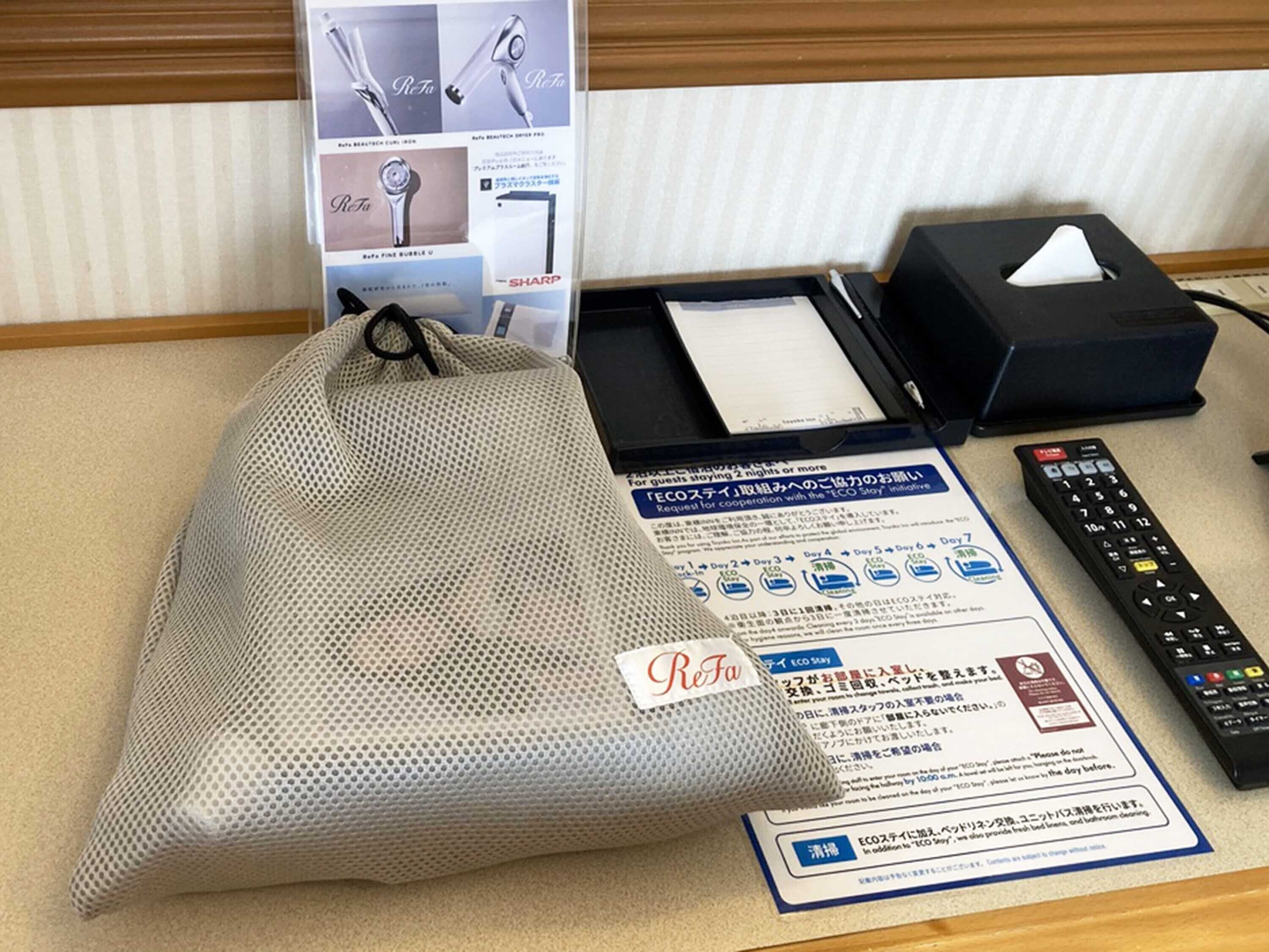 A hotel room with a bed, a remote control, a tissue box, and a ReFa branded bag.