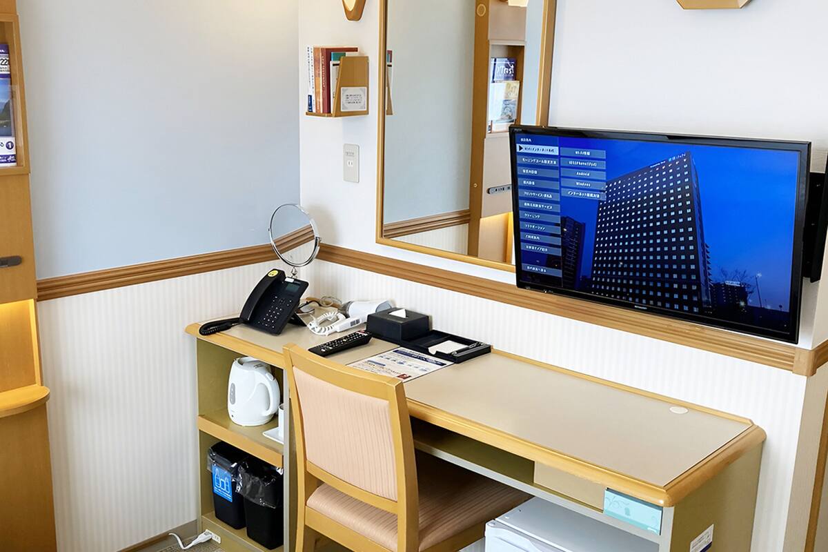 A hotel room with a desk, a telephone, a computer monitor, a mirror, and a small refrigerator.