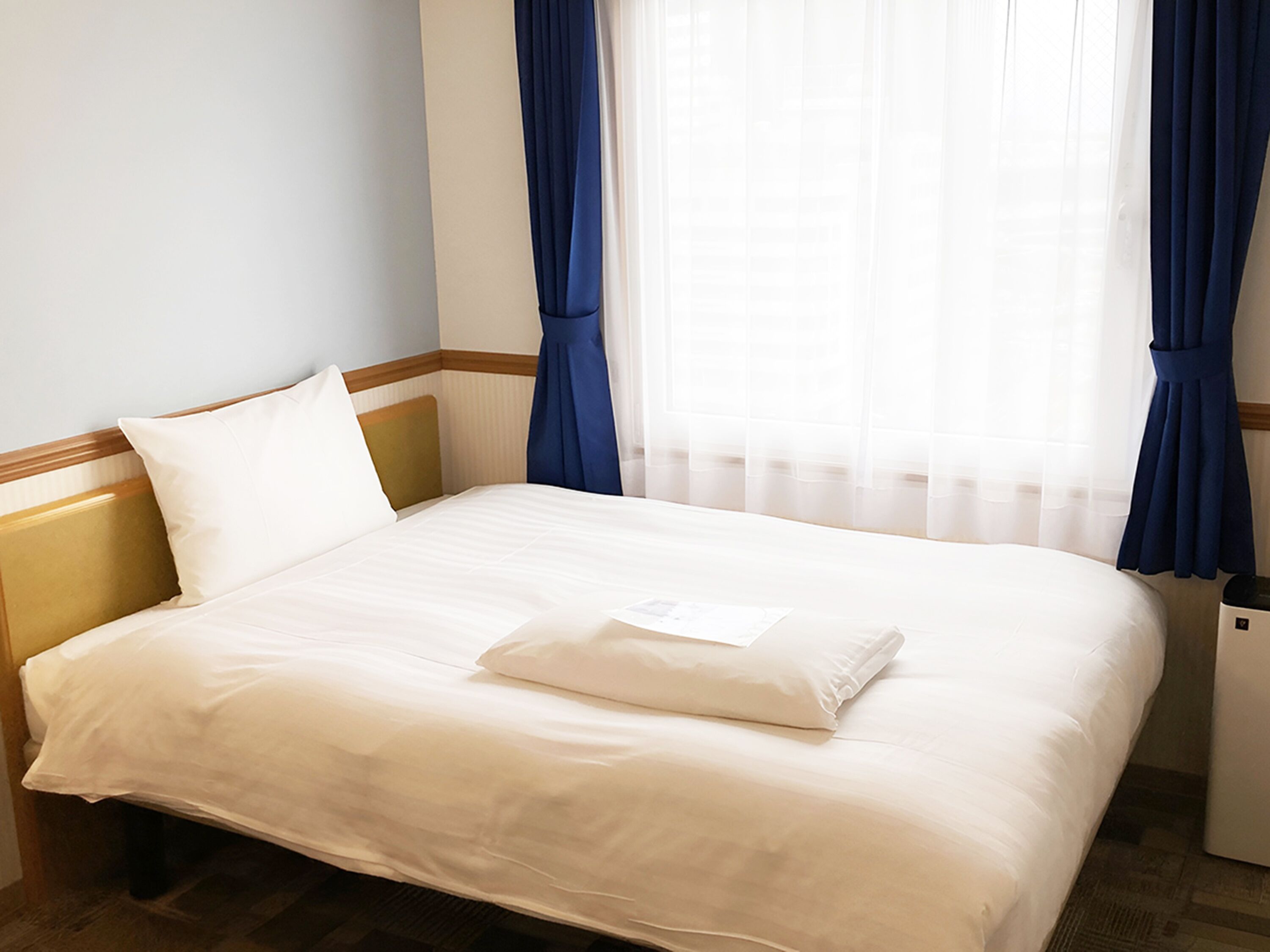 Een net opgemaakt bed met witte lakens in een hotelkamer, met een houten hoofdeinde, een klein nachtkastje en blauwe gordijnen.