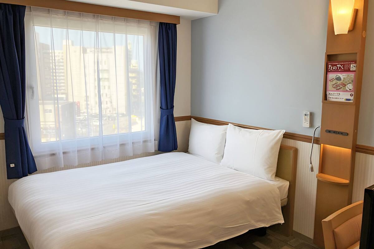 A hotel room with a large bed, a window with curtains, a bedside table, and a wall-mounted light fixture.