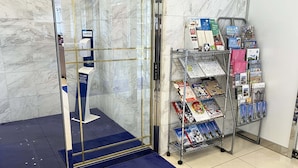 A reception area with a marble wall, a glass elevator, a blue carpet, and a magazine rack with various publications.
