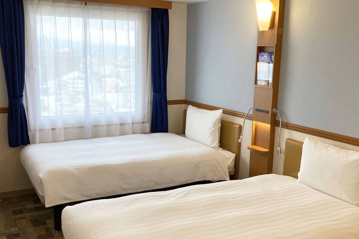 A hotel room with two beds, a window with sheer curtains, a bedside table with a lamp, and a wall-mounted sign.
