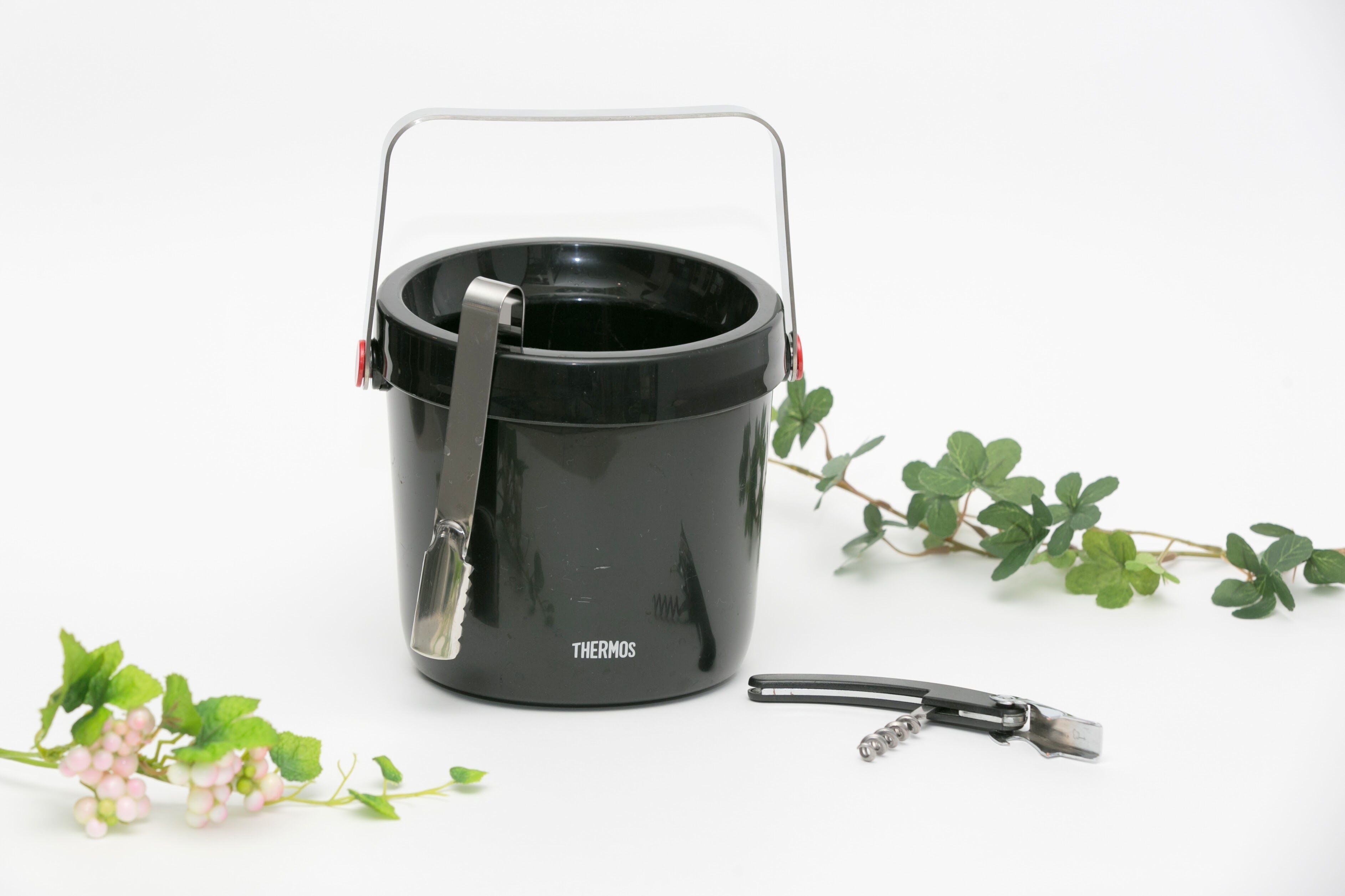 A black thermos with a handle and a strap, placed on a white surface with green foliage.