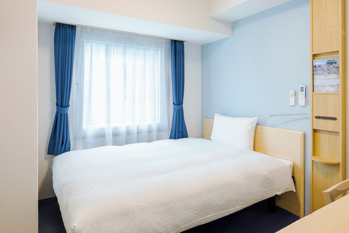A hotel room with a large bed, a bedside table, a wooden wardrobe, and a window with curtains.