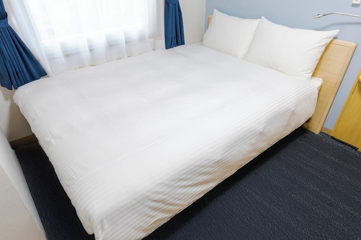 A neatly made bed with white linens in a room with a dark carpet, a wooden headboard, and a window with sheer curtains.