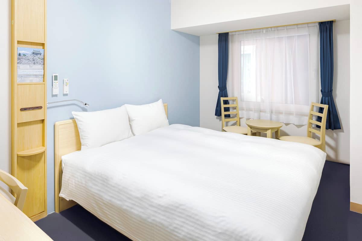 A hotel room with a large bed, two chairs, a small table, and a window with curtains.