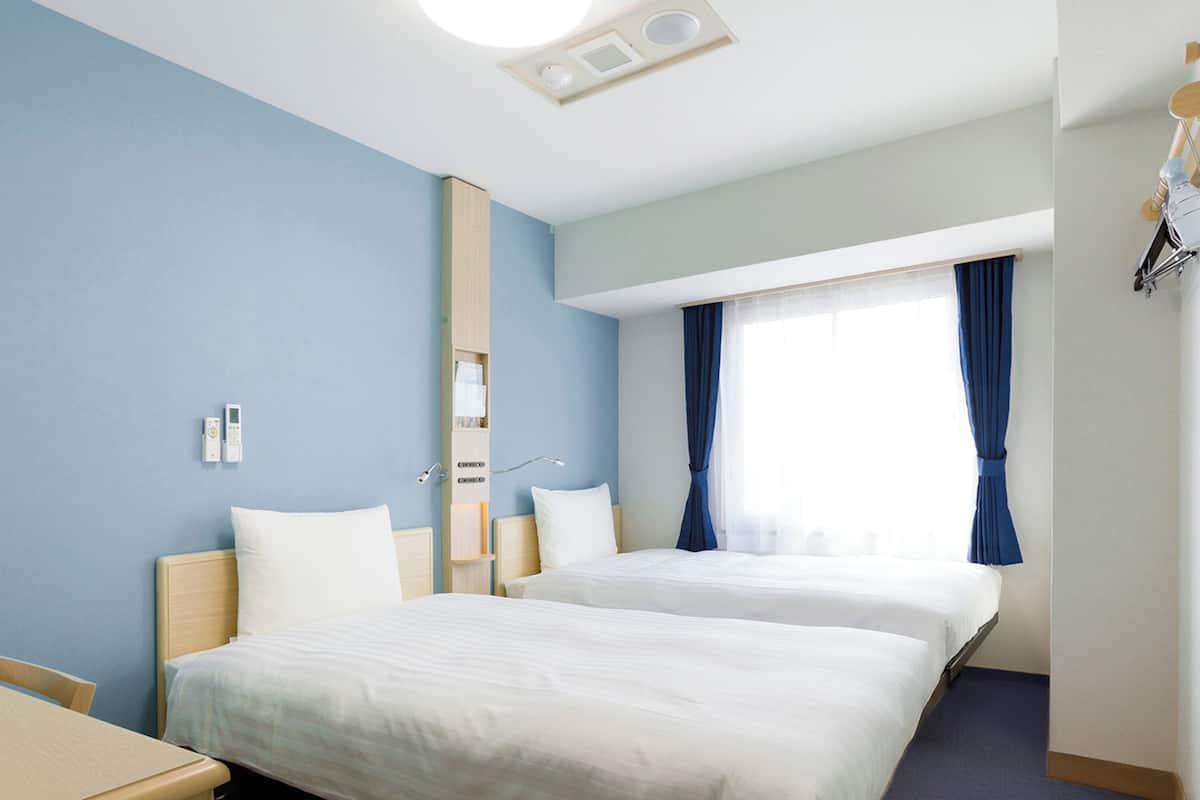 A hotel room with two beds, a bedside table, a nightstand, a window with curtains, and a ceiling light.