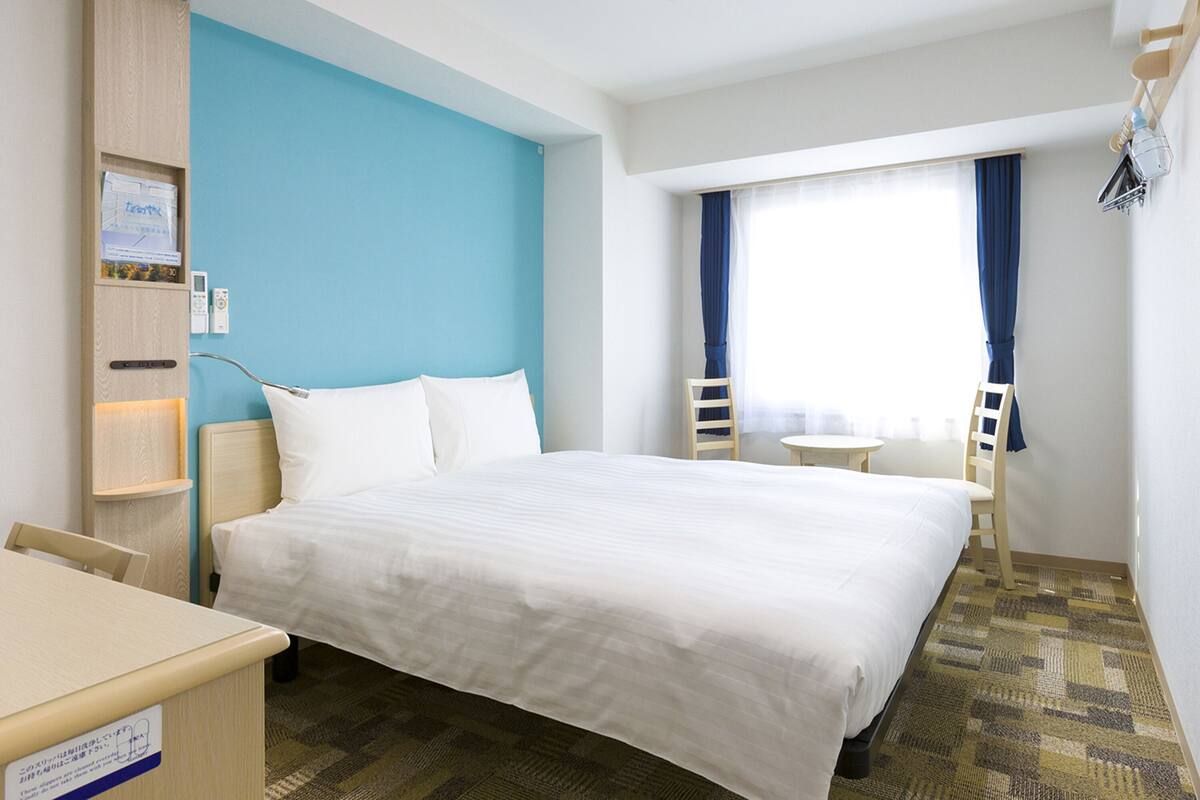 Ein Hotelzimmer mit Bett, Schreibtisch, Stuhl, kleinem Tisch und einem Fenster mit Vorhängen.