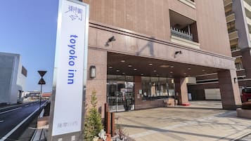 The exterior of the Toyoko Inn, featuring a prominent entrance with a clear pathway leading to the main doors, a visible parking area, and a sign indicating the hotel's name.