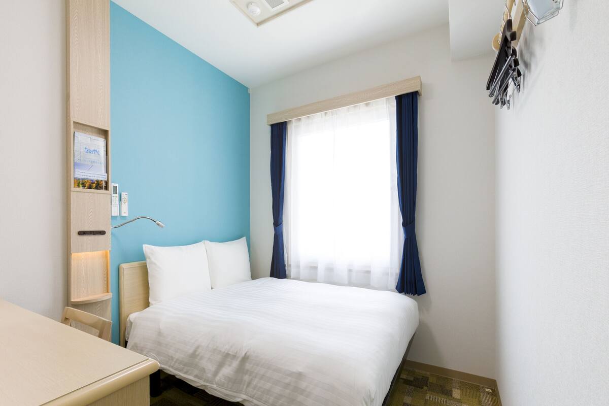 Ein Hotelzimmer mit einem Bett, einem Schreibtisch, einem Nachttisch, einem an der Wand befestigten Ablagefach mit Toilettenartikeln und einem Fenster mit Vorhängen.