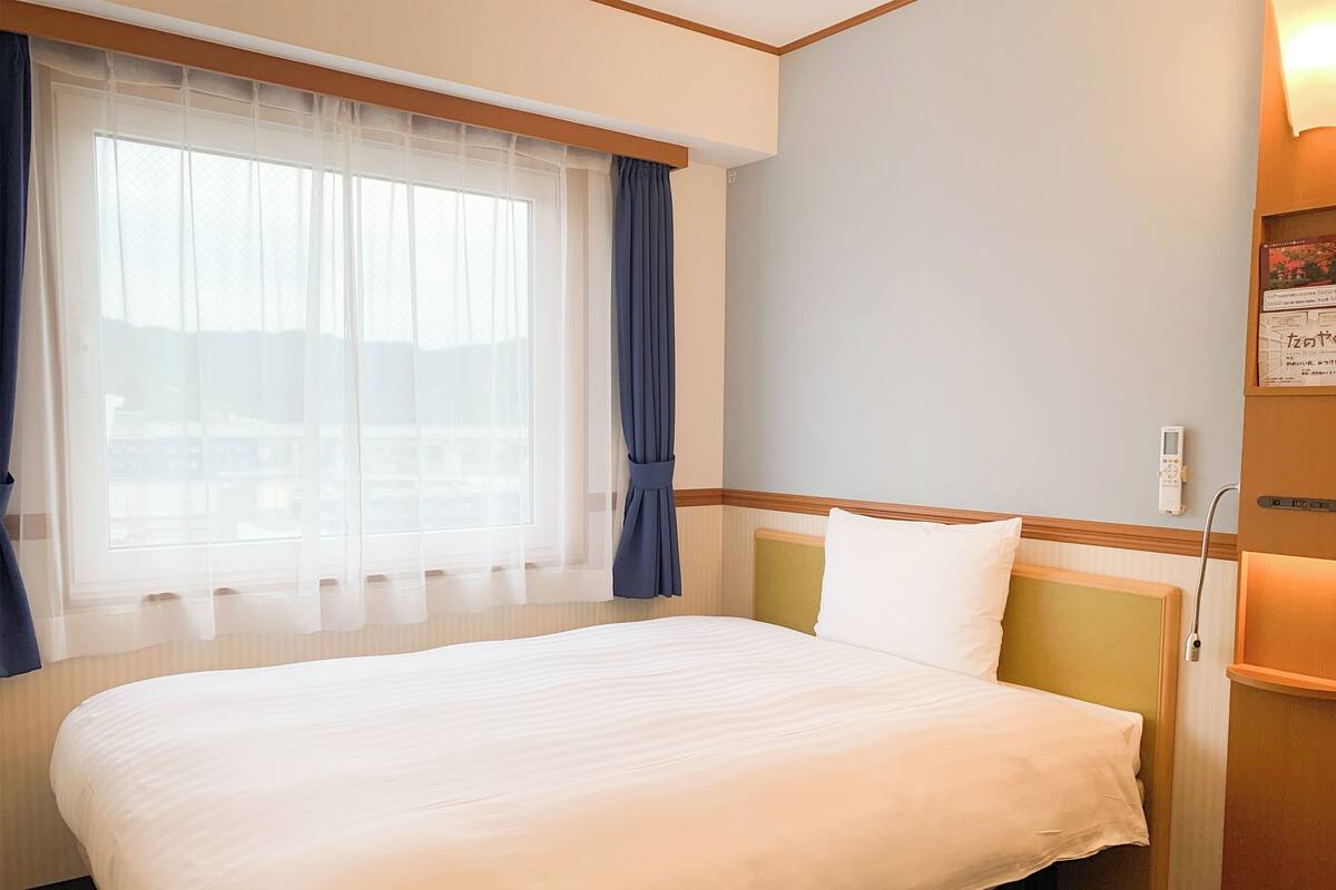 A hotel room with a large bed, a window with sheer curtains, a bedside table, and a wall-mounted control panel.