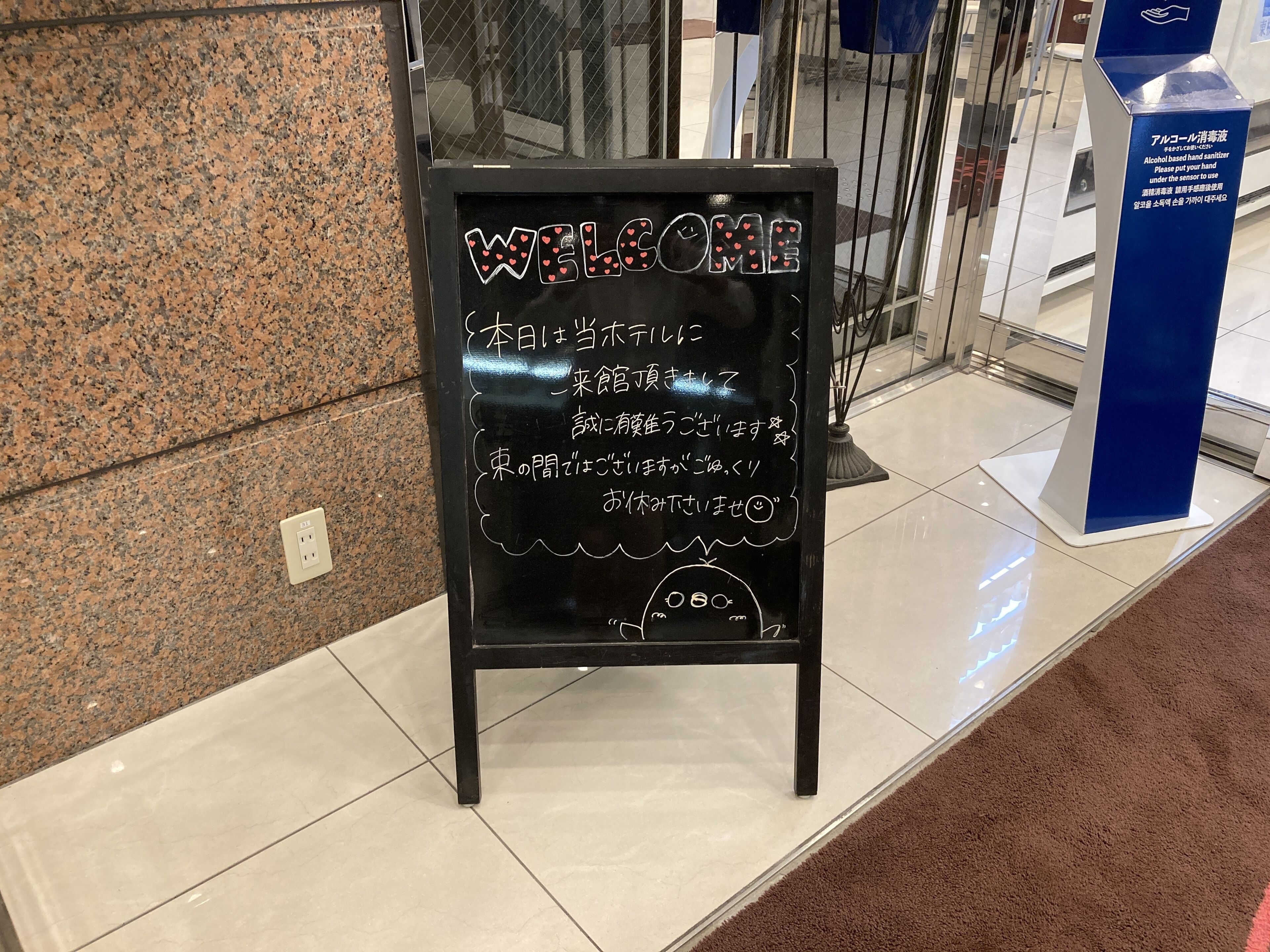 Una pizarra con un mensaje de bienvenida y texto en japonés, ubicada sobre un suelo de baldosas junto a una puerta de cristal.