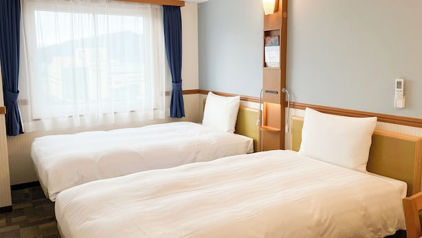 A hotel room with two beds, a window with curtains, a bedside table with a lamp, and a thermostat on the wall. - Toyoko Inn Tsuruga Ekimae (Tsuruga)