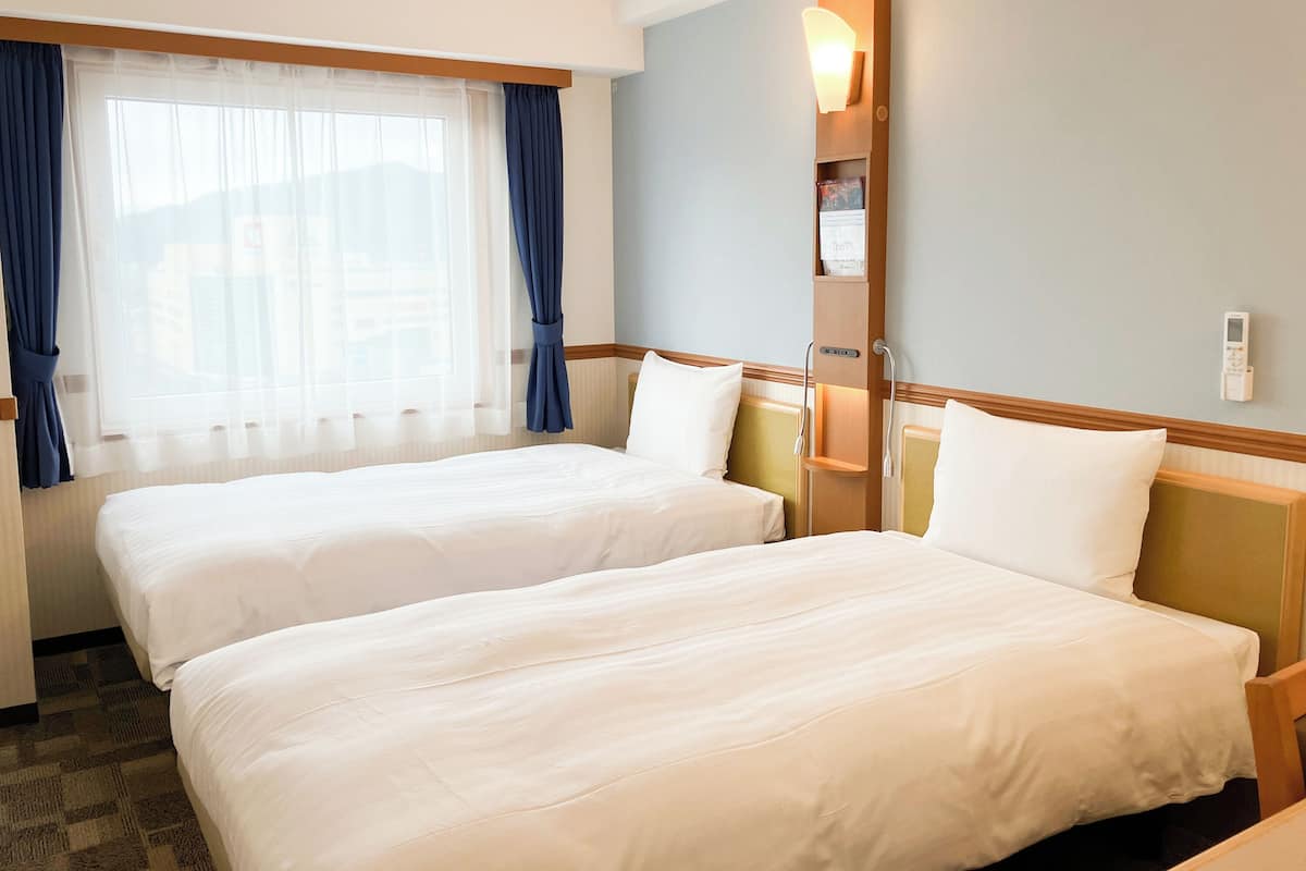 A hotel room with two beds, a window with curtains, a bedside table with a lamp, and a thermostat on the wall.