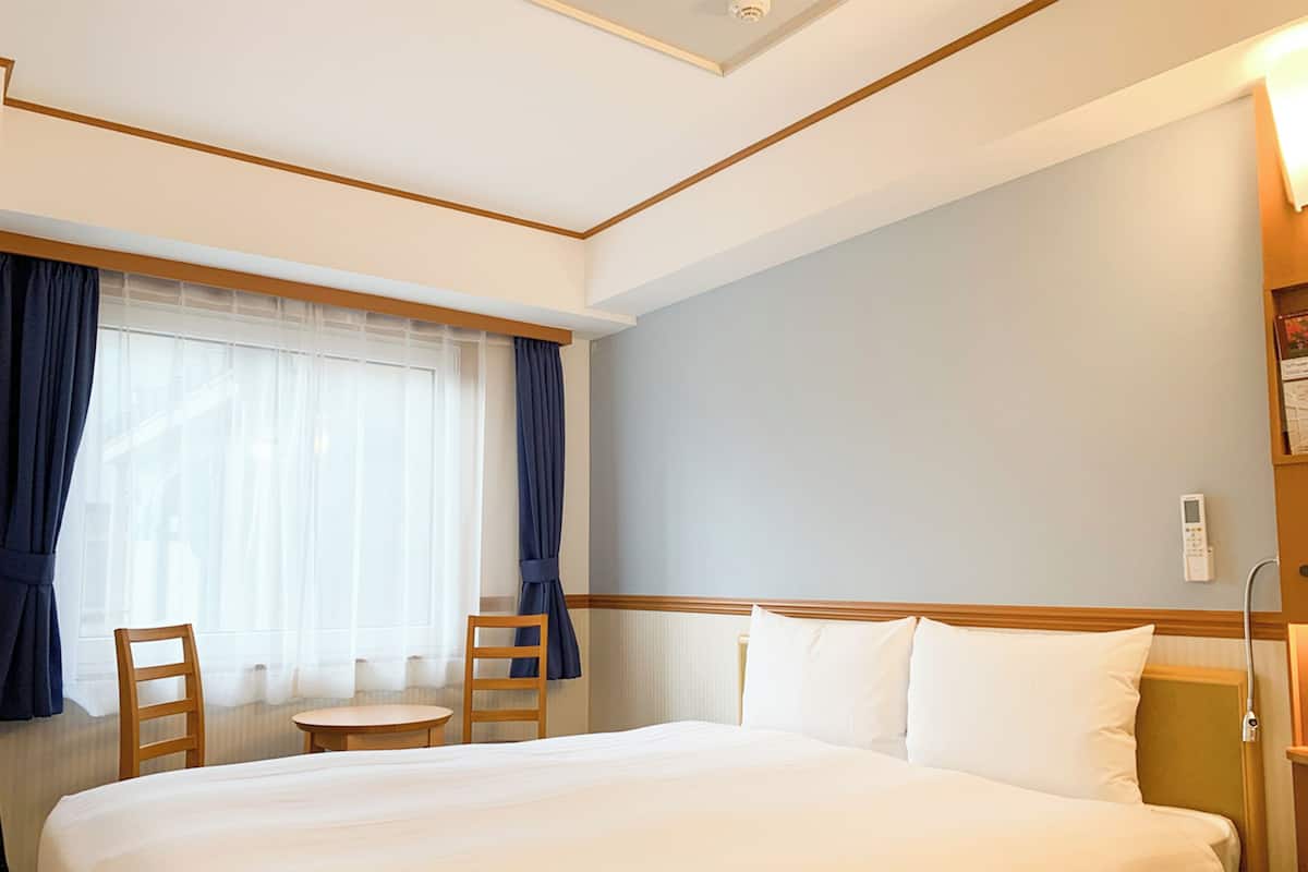 A hotel room with a large bed, a small table, a chair, and a window with curtains.