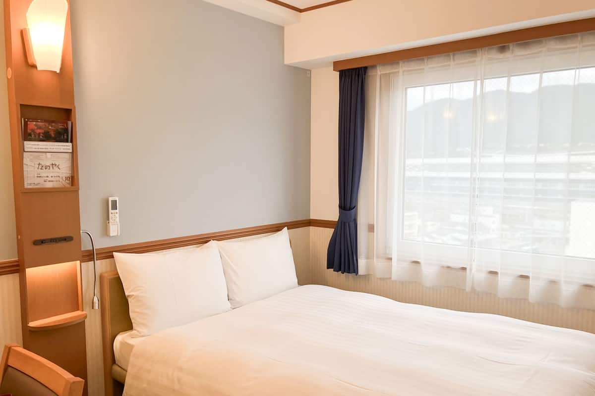 A hotel room with a large bed, a bedside table, a chair, a window with curtains, and a wall-mounted light fixture.