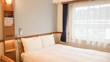 A hotel room with a large bed, a bedside table, a chair, a window with curtains, and a wall-mounted light fixture.