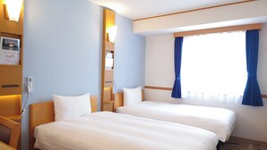 Habitación de hotel con dos camas, una mesita de noche, repisa con lámpara y ventana con cortinas.