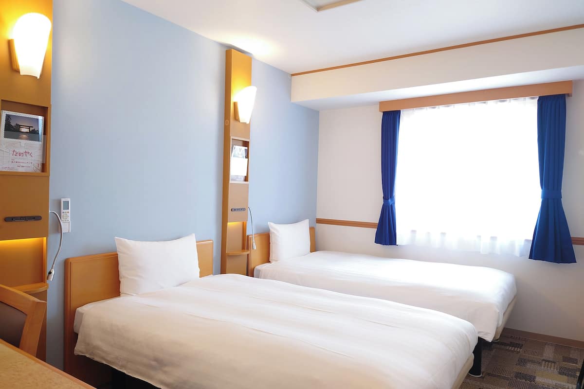A hotel room with two beds, a bedside table, a wall-mounted shelf with a lamp, and a window with curtains.