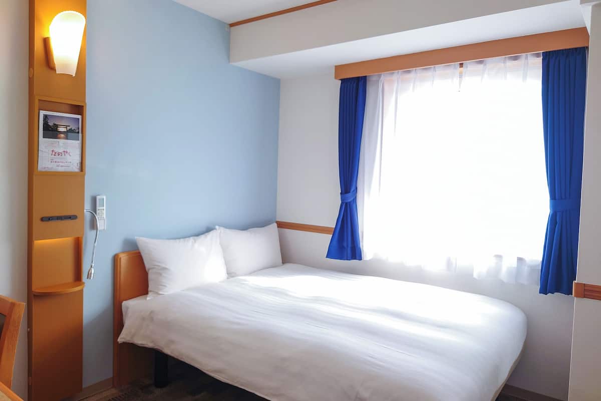 A hotel room with a bed, a nightstand, a wall-mounted lamp, and a window with curtains.