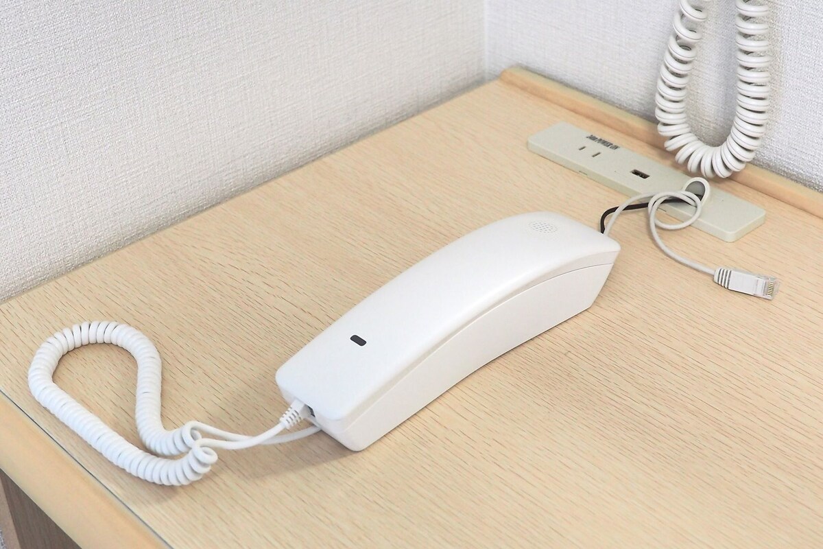 Teléfono blanco sobre una mesa de madera con un cable enrollado, un adaptador de corriente y un enchufe de pared.