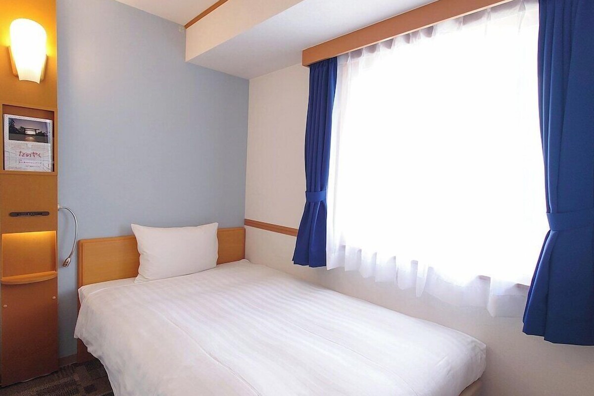 A hotel room with a bed, a nightstand, a wall-mounted lamp, and a window with curtains.