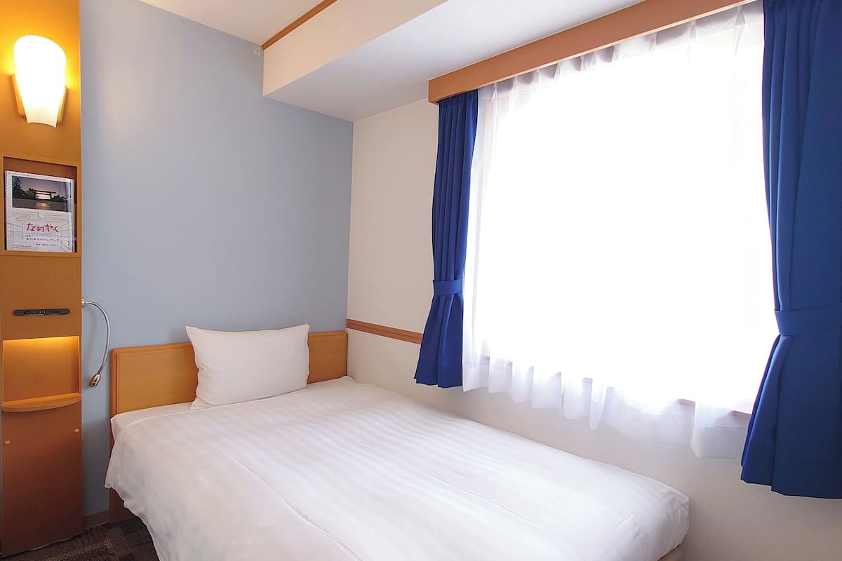 A hotel room with a bed, a nightstand, a wall-mounted lamp, and a window with curtains.