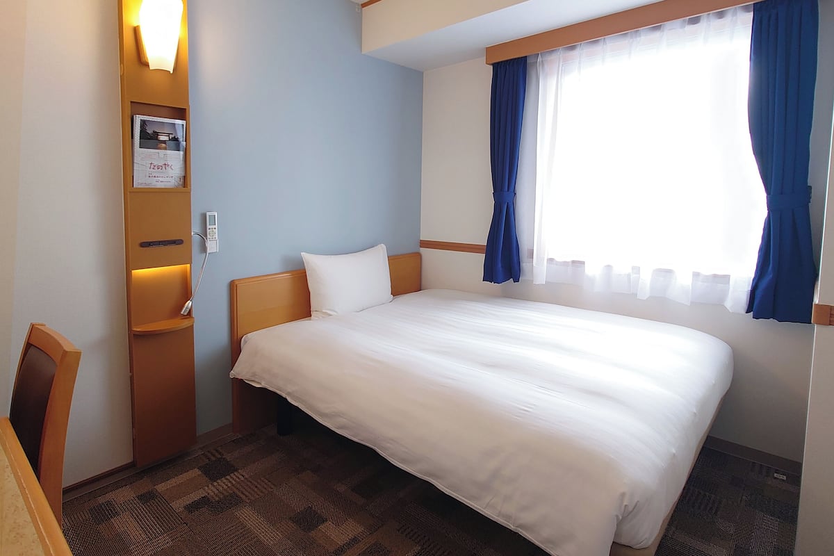 A hotel room with a large bed, a bedside table, a lamp, a wall-mounted thermostat, and a window with curtains.