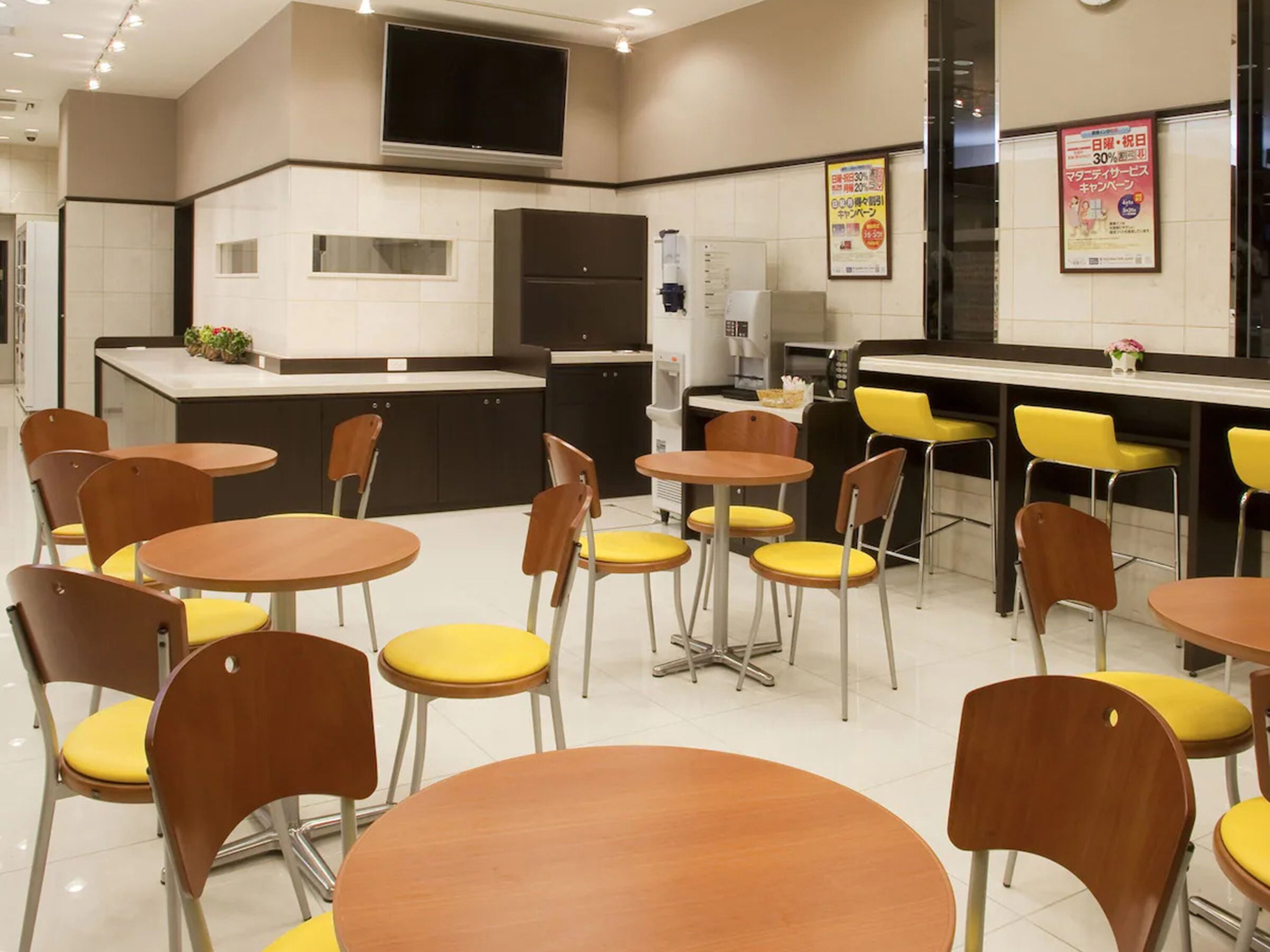 A modern kitchen with a flat-screen TV, a coffee machine, and a refrigerator. There are several yellow and brown chairs around small tables.