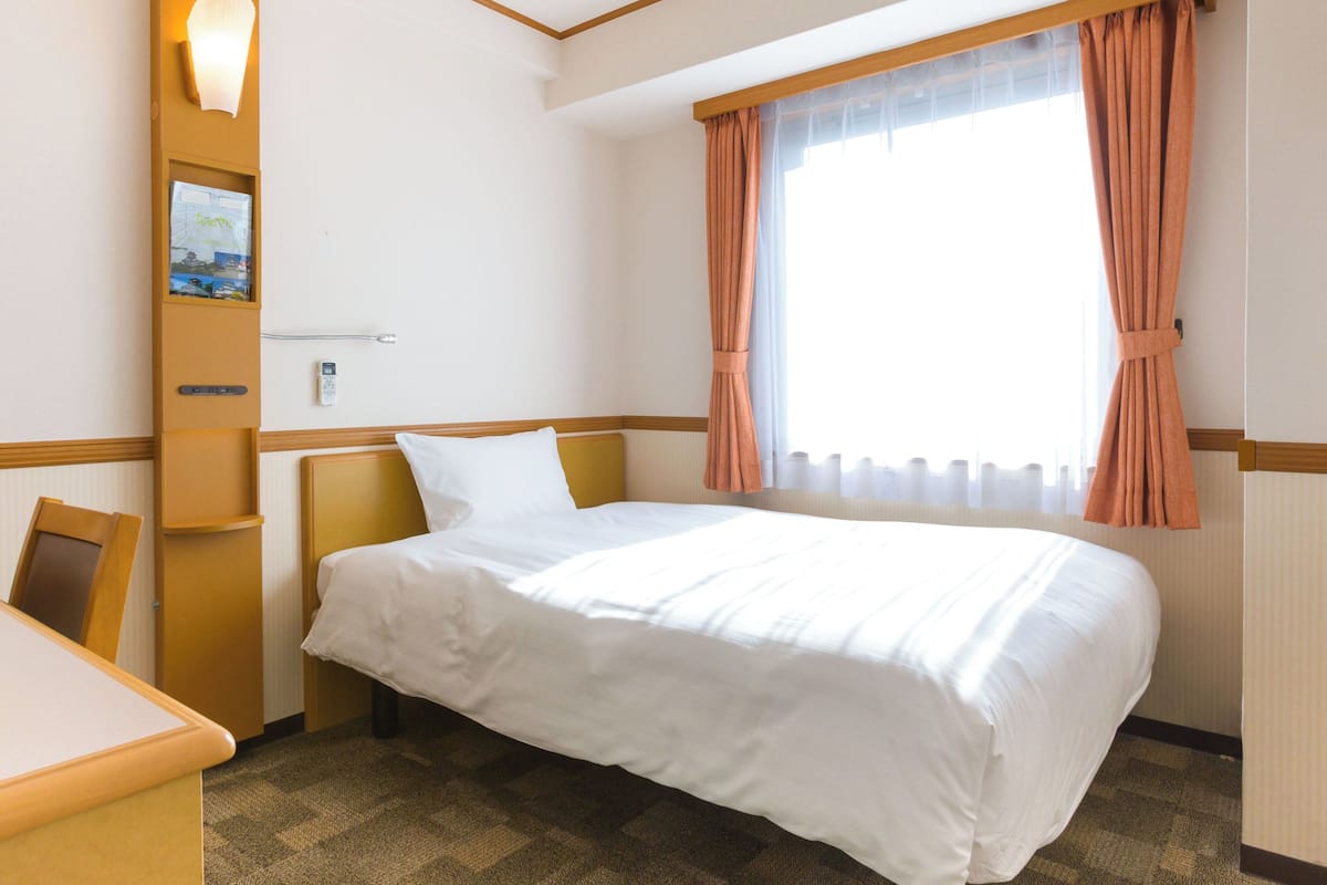 Ein Hotelzimmer mit Bett, Schreibtisch, Stuhl, einer Wandablage mit Lampe und einem fenster mit Vorhängen.