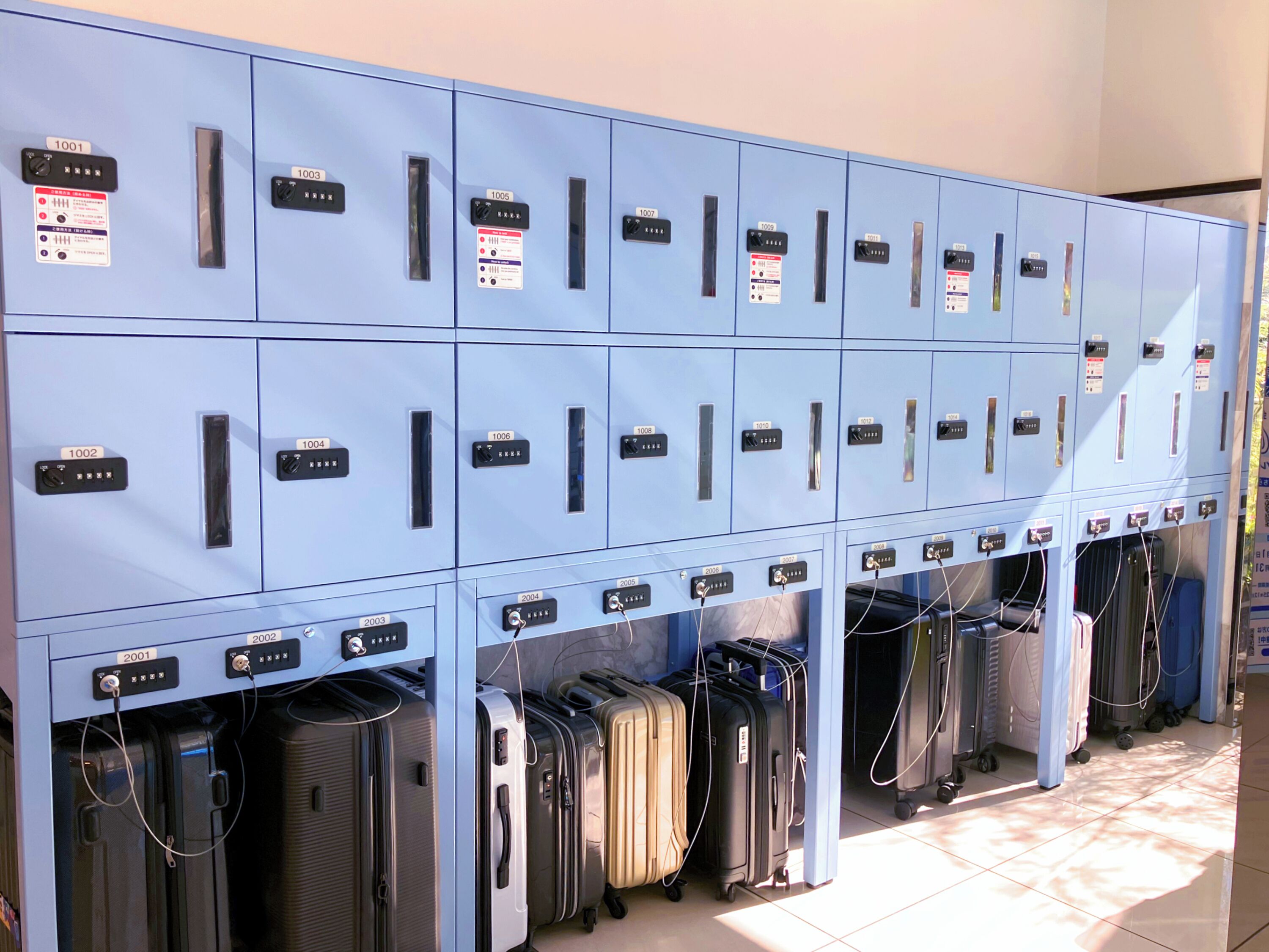 Een rij blauwe lockers met genummerde labels, waarvan enkele elektronische sloten hebben, en diverse koffers eronder.