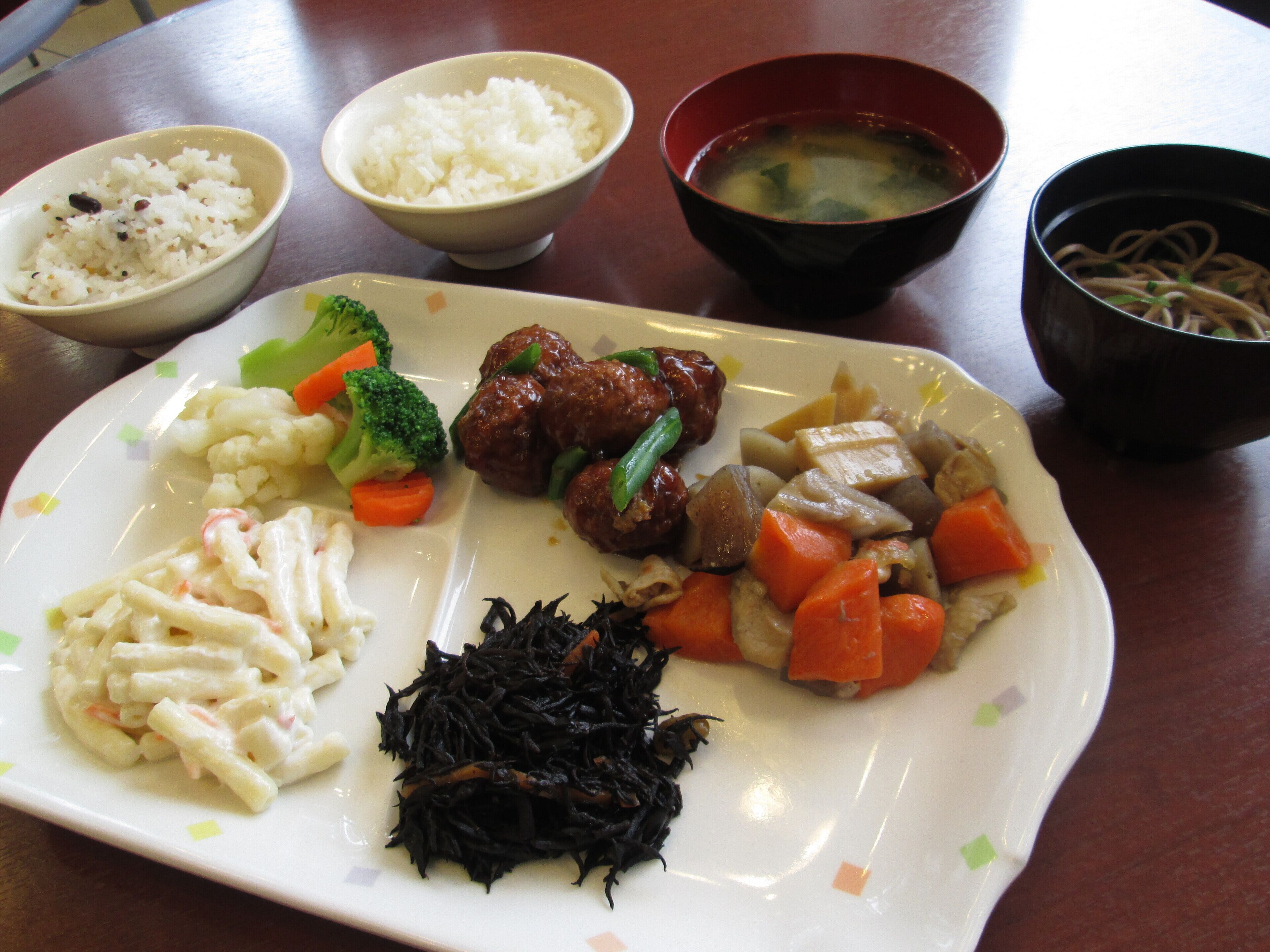 Une assiette contenant divers plats, notamment du riz, de la soupe, de la viande, des légumes et des nouilles.