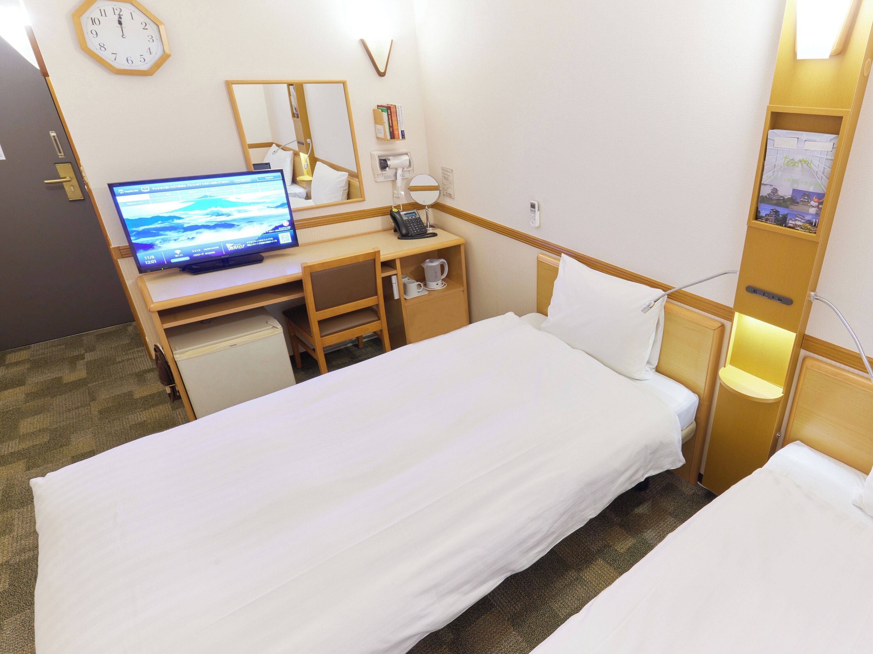A hotel room with a bed, a desk with a computer, a chair, a small refrigerator, a bedside table with a lamp, and a wall clock.