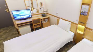 A hotel room with a bed, a desk with a computer, a chair, a small refrigerator, a bedside table with a lamp, and a wall clock.
